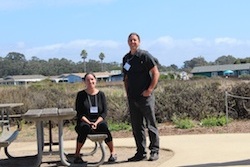 two people outside, one sitting on a picnic table while the other stands